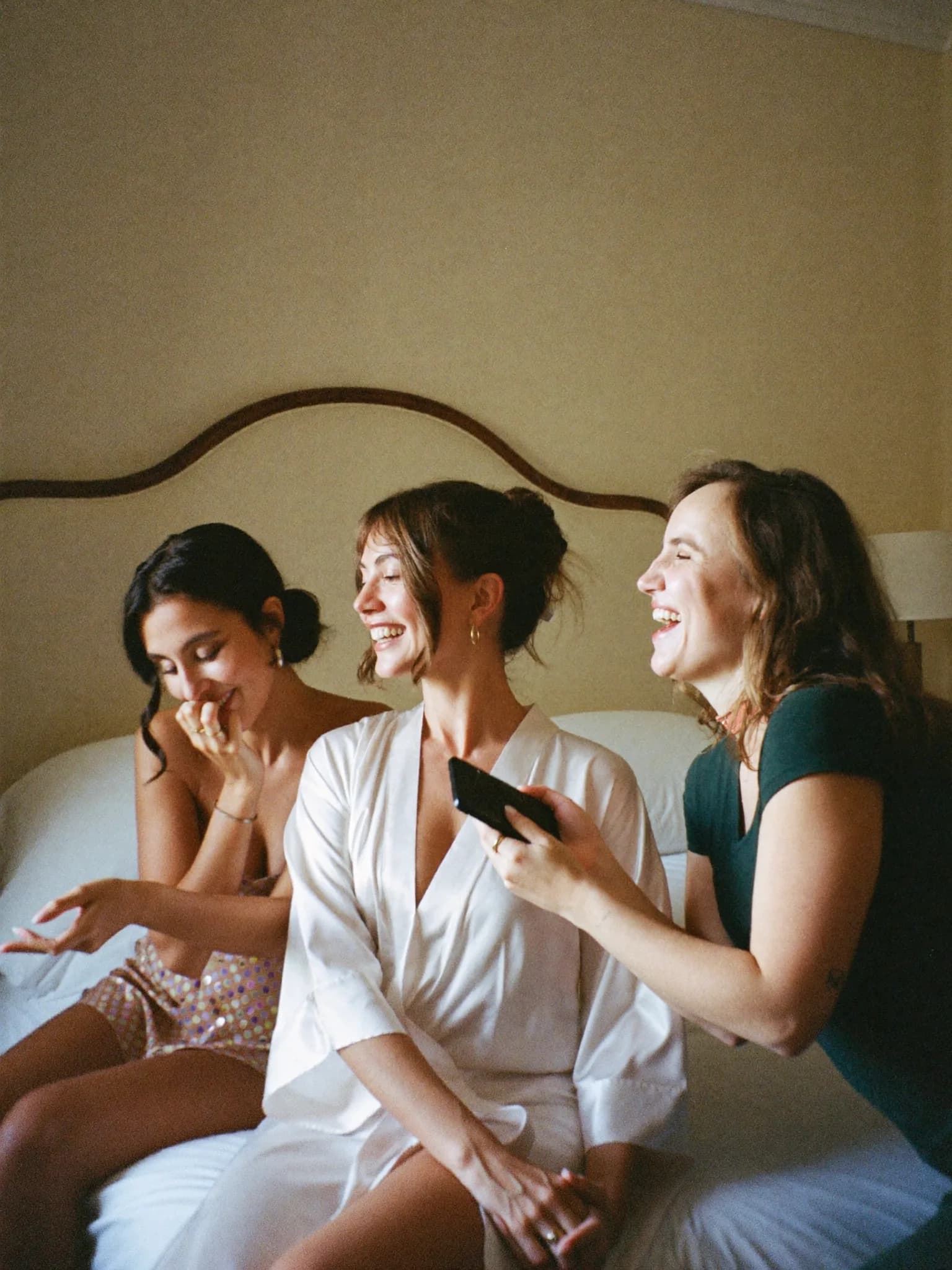 Generata con IA: una sposa in vestaglia di seta ride con due amiche, una le mostra qualcosa sul telefono.